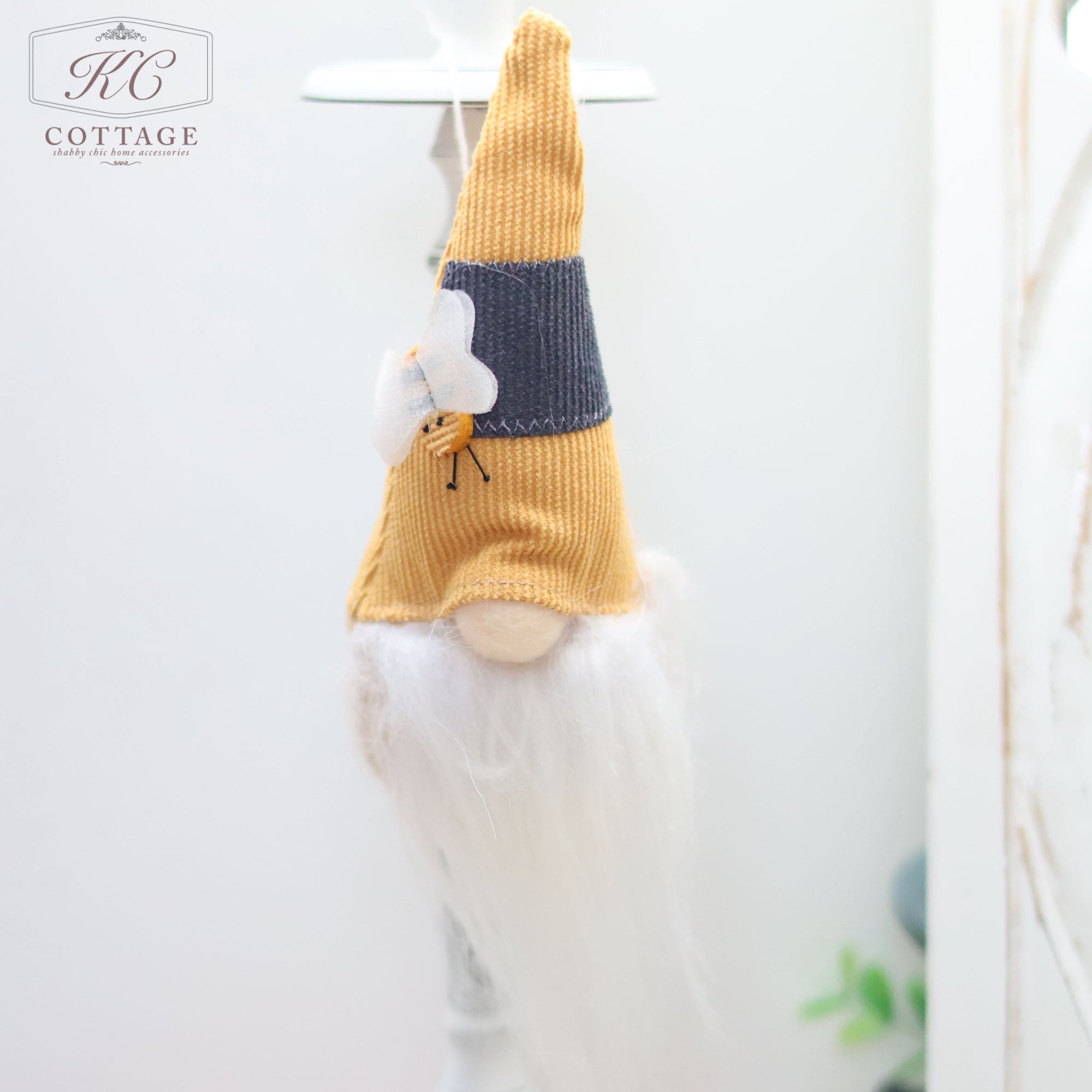 A Fabric Hanging Bee Gonk with a long white beard, yellow hat, and grey patch hangs from a white hook. The gnome has an orange body and a small white butterfly attached to its hat. Perfect for enhancing your home decor, the setting is bright and minimal.