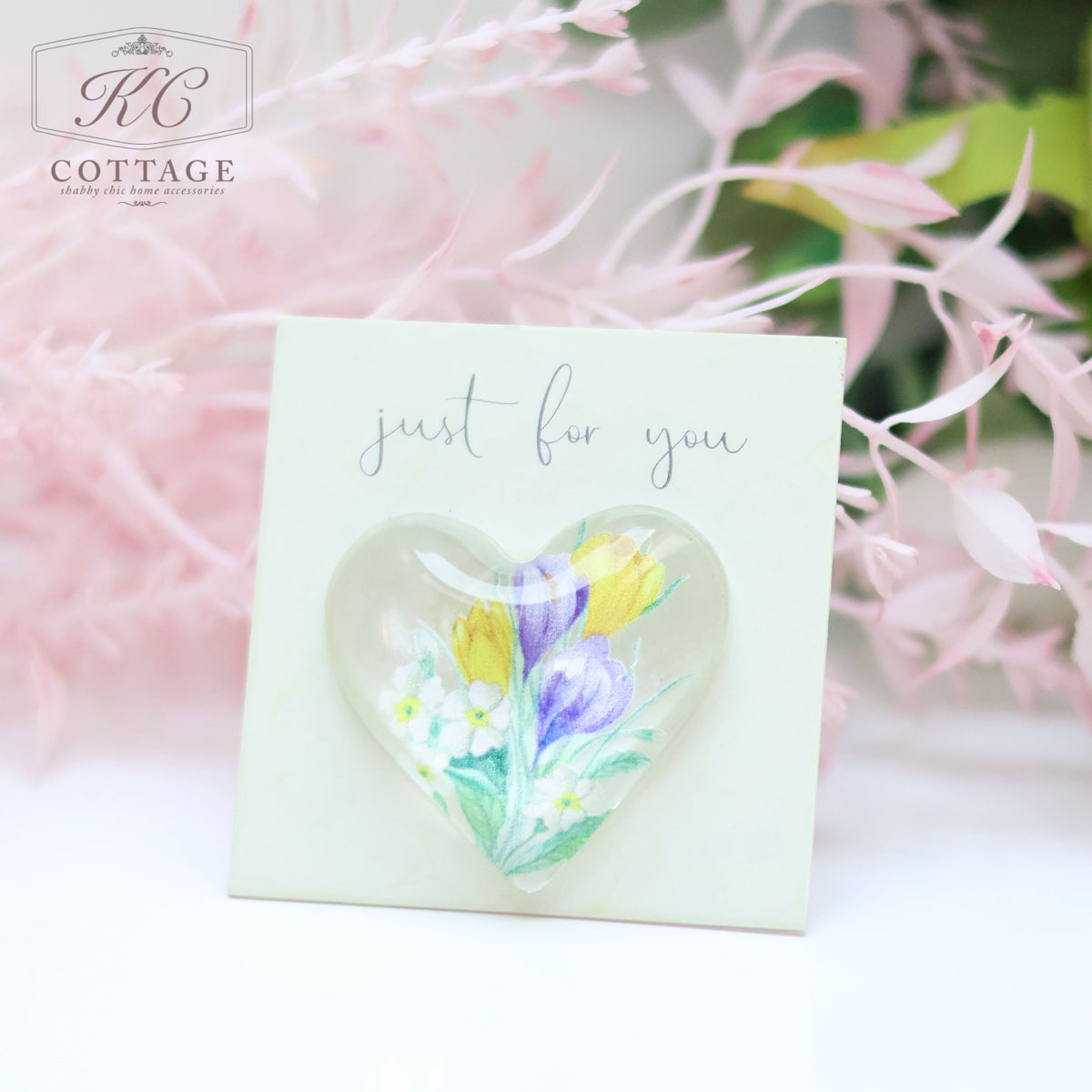 A Glass Spring Floral Heart with Sentiment Card with a colorful floral design is displayed on a small square card with the text "just for you." The card is positioned in front of a soft blurred background featuring pink and green foliage. Perfect for adding charm to your home decor.