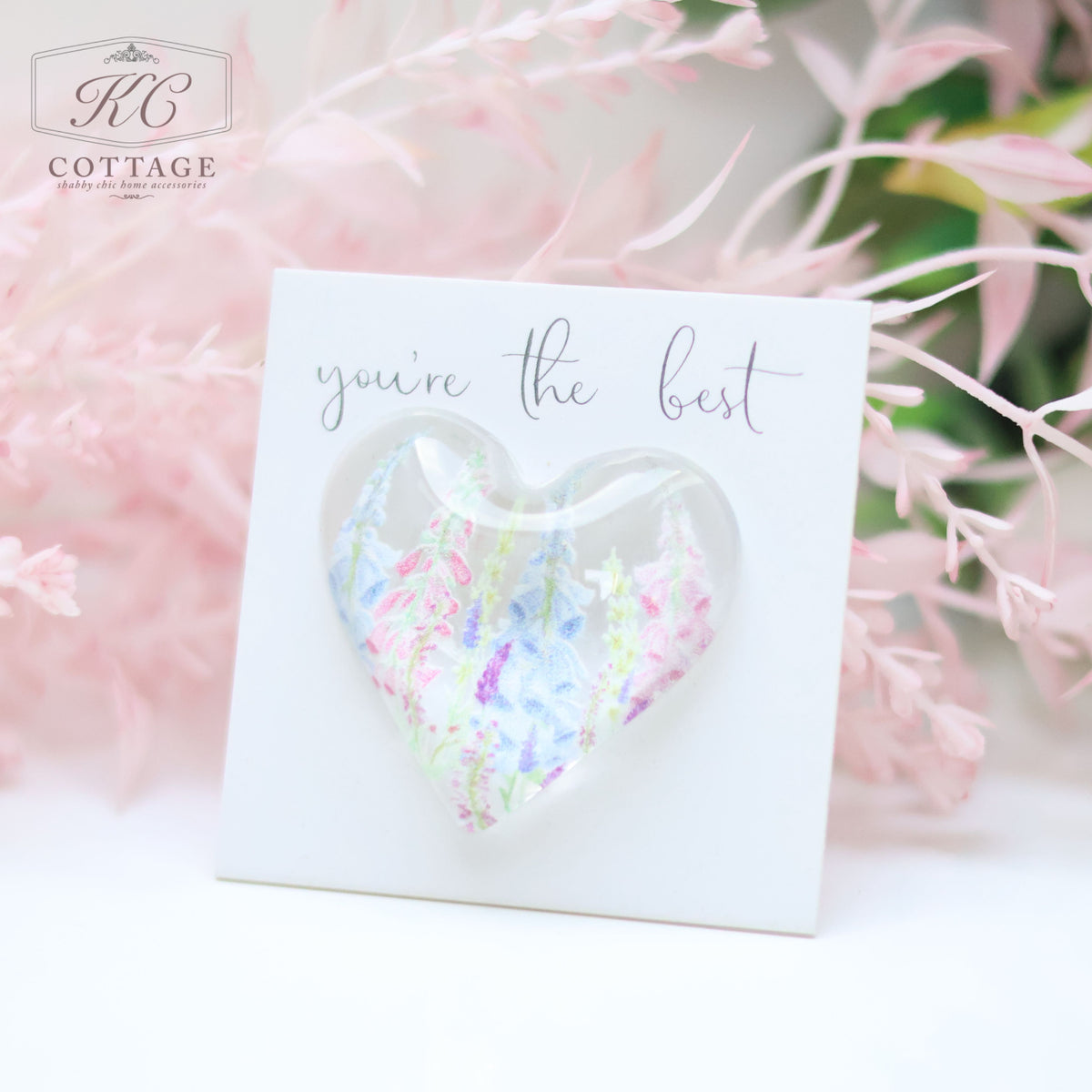 A decorative card with a white background displays a heart-shaped clear bubble containing pastel-colored wildflowers. The card reads "you're the best." Pink and green foliage is blurred in the background, making it an ideal piece of home decor. This Glass Spring Floral Heart with Sentiment Card is perfect for adding a touch of charm to any space.