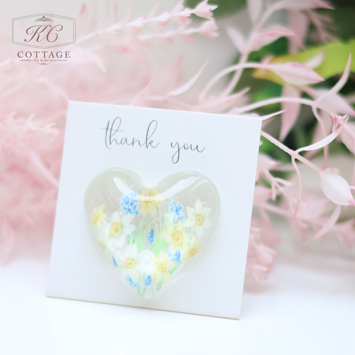 A Glass Spring Floral Heart with Sentiment Card is mounted on a white card with "thank you" written in cursive. The card is set against a background of soft pink and green flowers, perfect for adding a touch of elegance to your home decor.