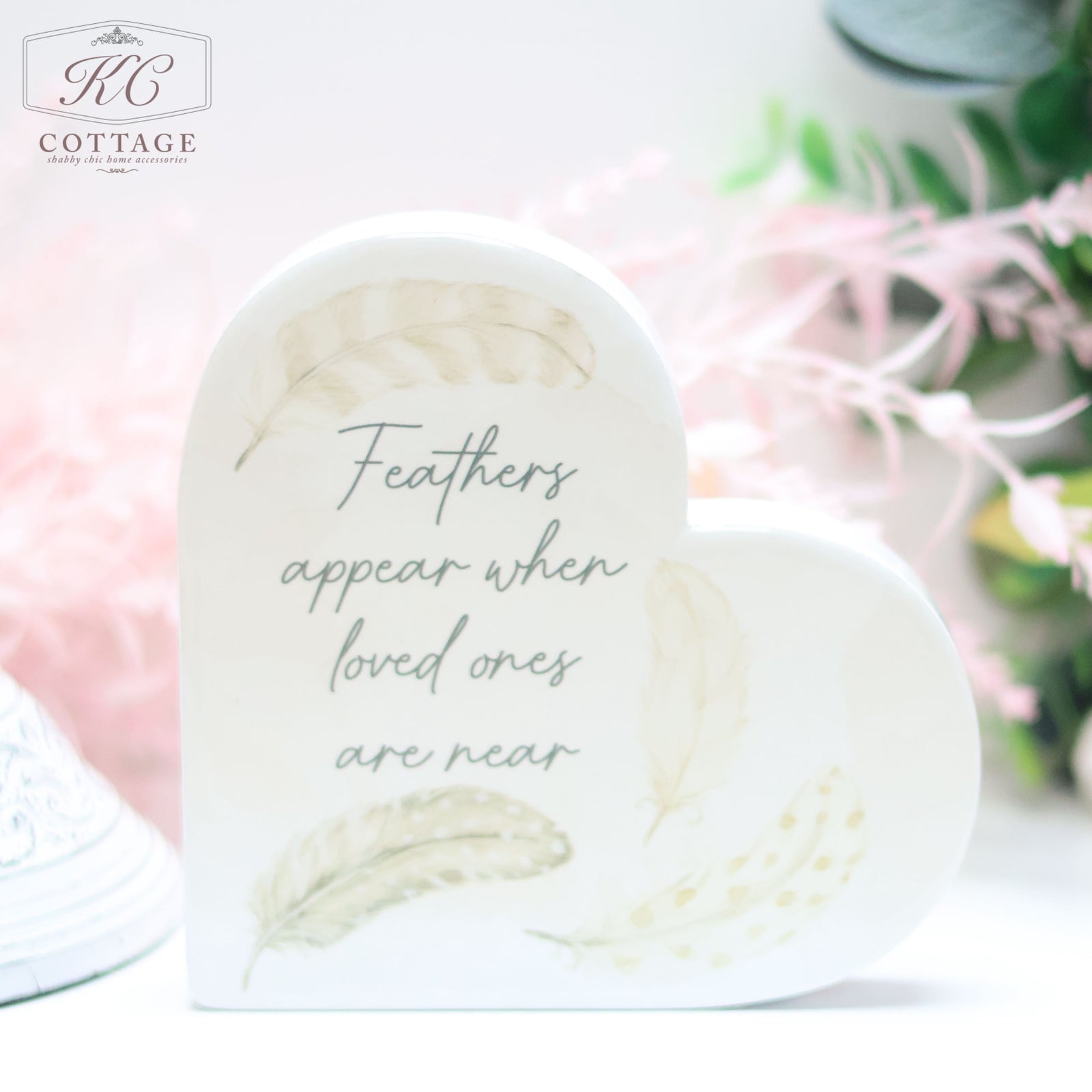 A Grey Feather Ceramic Memory Heart Block with light feather illustrations and the text "Feathers appear when loved ones are near" is displayed in soft focus. The background includes a pink, green, and white floral arrangement, making it perfect for home decor enthusiasts.