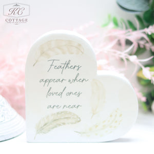 A Grey Feather Ceramic Memory Heart Block with light feather illustrations and the text "Feathers appear when loved ones are near" is displayed in soft focus. The background includes a pink, green, and white floral arrangement, making it perfect for home decor enthusiasts.