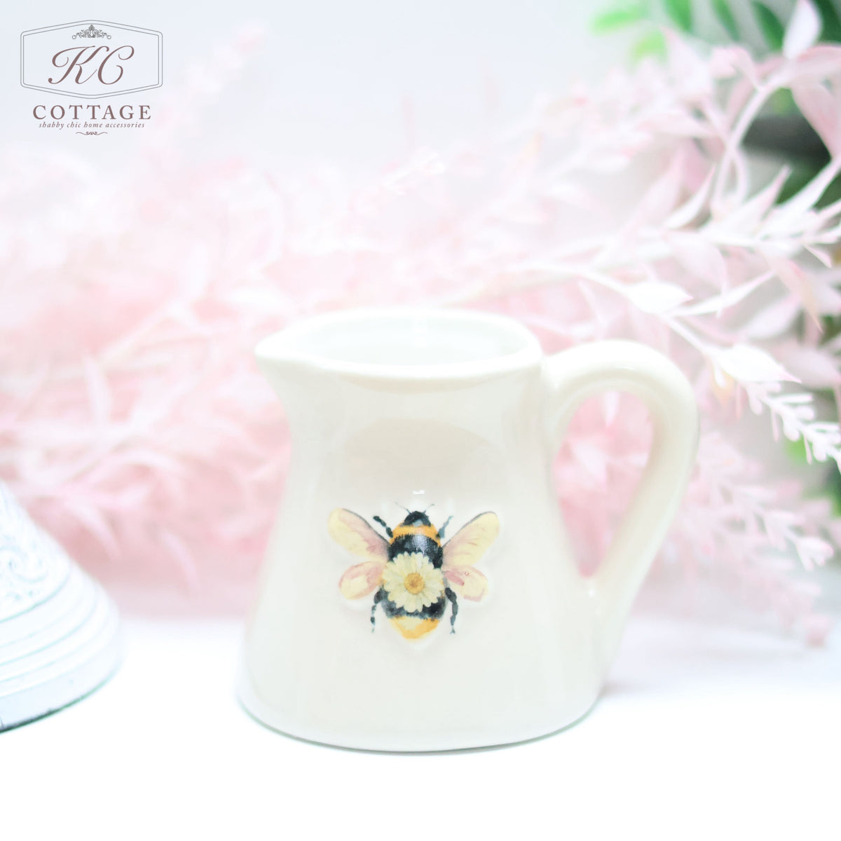 A small white Daisy Bee Mini Milk Jug with a colorful bee illustration on its front is placed against a soft pink floral background. The pitcher, ideal for home decor, has a handle and an open top.