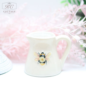 A small white Daisy Bee Mini Milk Jug with a colorful bee illustration on its front is placed against a soft pink floral background. The pitcher, ideal for home decor, has a handle and an open top.
