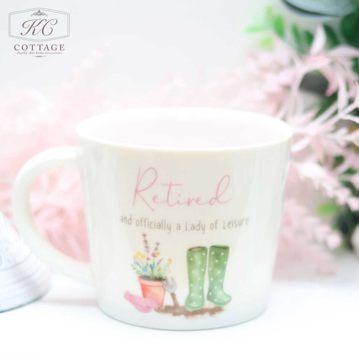 A Retirement Mug With Wellies & Plant Pot with the text "Retired and officially a Lady of Leisure" in pink and gray. The mug has illustrations of green polka dot boots, a watering can, a flower pot, and flowers. Perfect for adding charm to your home decor, the background features a blurred, pastel-toned floral arrangement.