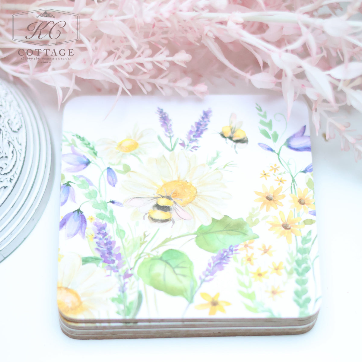 A Daisy Bee Floral Coaster featuring a watercolor-style illustration of various flowers, including daisies and lavender, with bees hovering over them. The background includes delicate, pink foliage. Perfect for home decor, the top left corner displays the branding "KC Cottage.