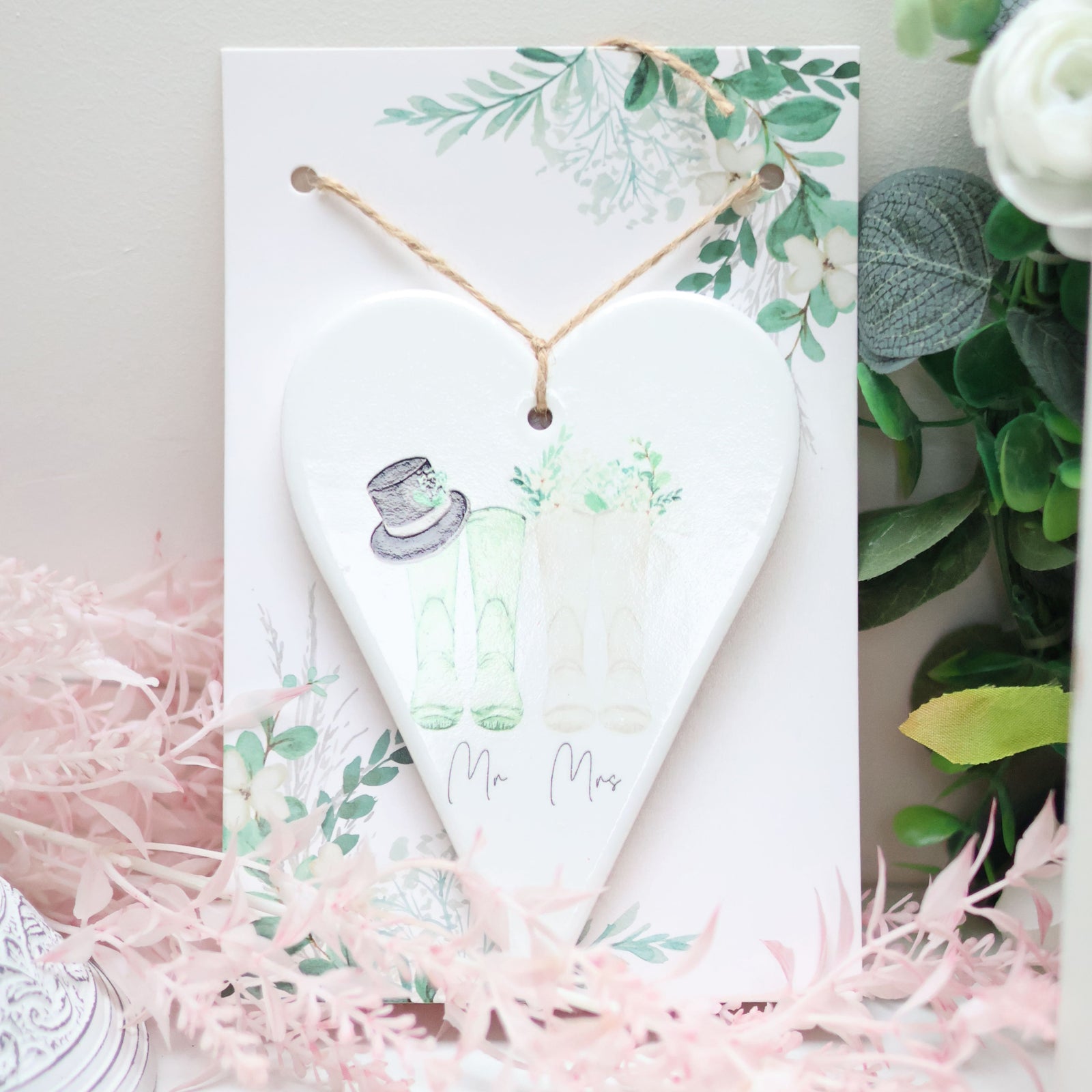 A Ceramic Wedding Heart with "Mr & Mrs" text hangs from a button-tied string. The home decor piece displays sketches of wedding attire: a top hat and veil, with greenery accents. Propped up against a card with similar botanical designs, it sits charmingly among floral arrangements.