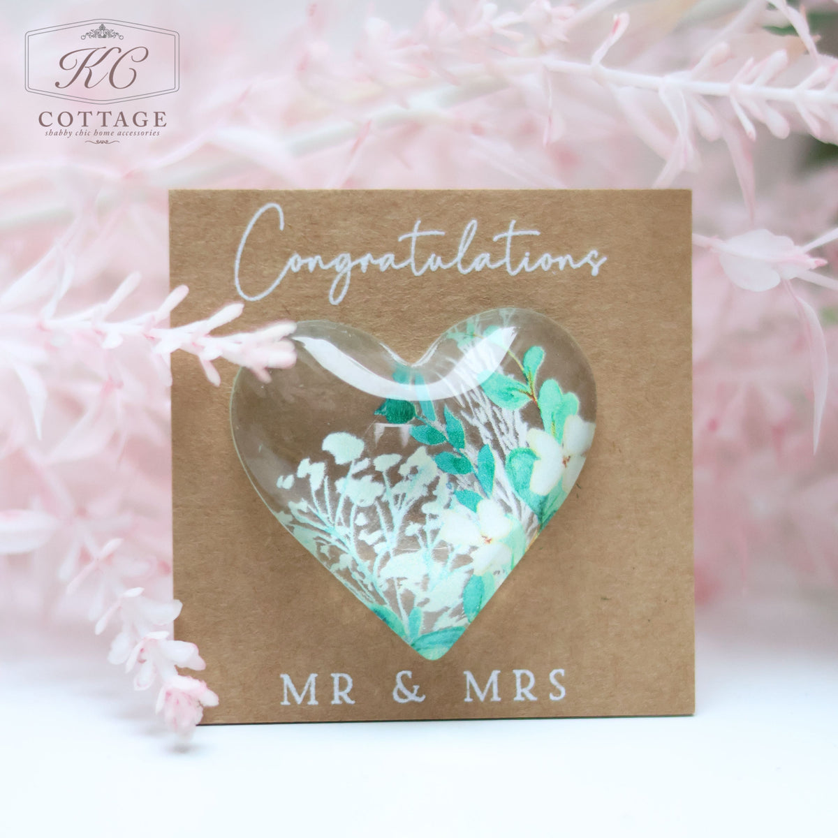 A **Wedding Pocket Heart Token On Card** with "Congratulations MR & MRS" written on it. The card features a heart-shaped window displaying a floral design with white and blue flowers. The background has delicate pink foliage, perfect for incorporating into your home decor.