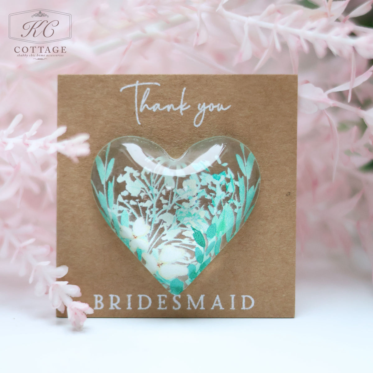 A small, heart-shaped Wedding Pocket Heart Token On Card featuring delicate white and teal flowers is attached to a brown card. The card has "Thank you" written at the top and "BRIDESMAID" at the bottom. Pink floral decorations surround the card, making it a charming piece of home decor.