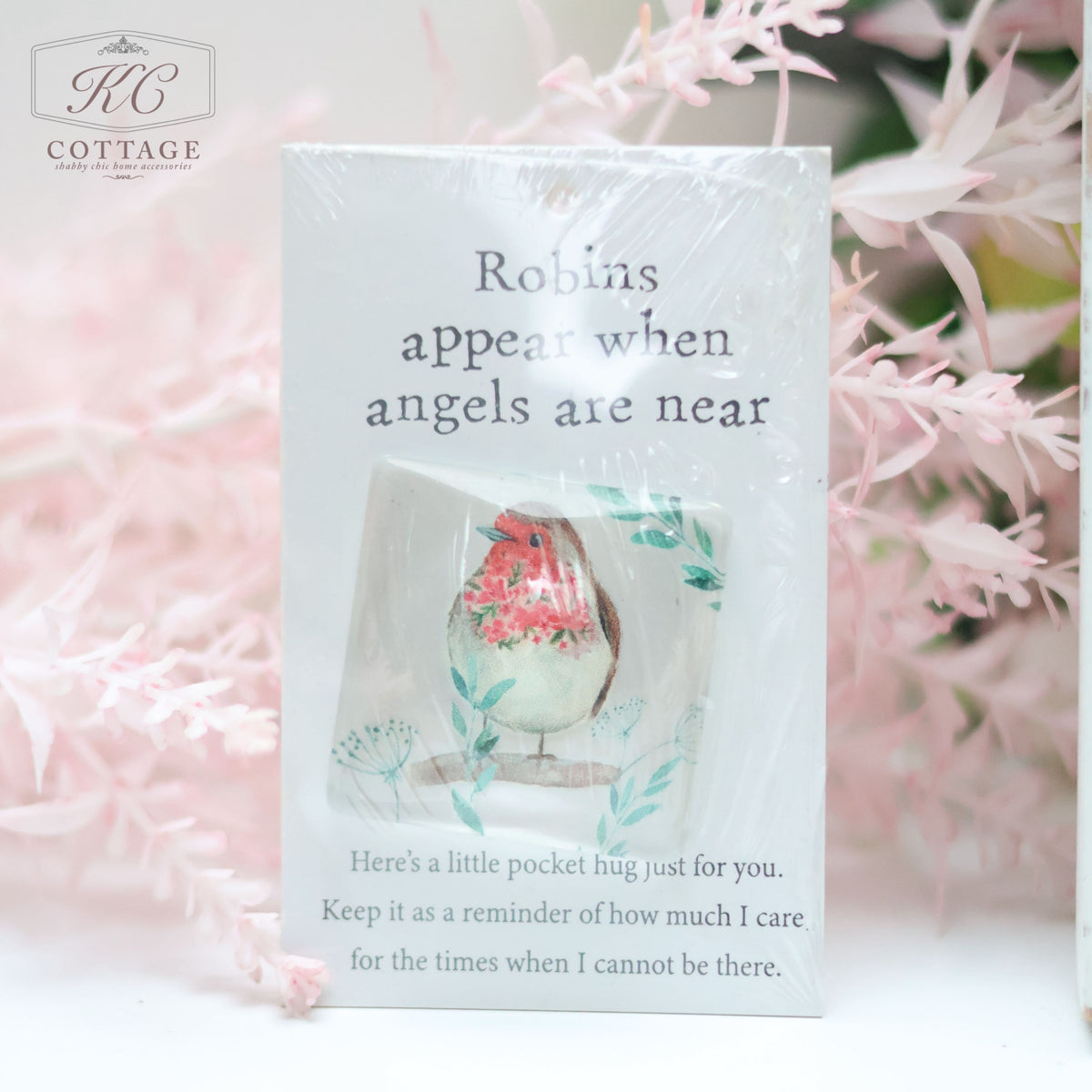 A small card with the text "Robins appear when angels are near" and an illustration of a robin. Below, it reads, "Here's a little pocket hug just for you. Keep it as a reminder of how much I care, for the times when I cannot be there." Surrounded by delicate pink foliage, Pocket Hugs with Sentiments is perfect for home decor.
