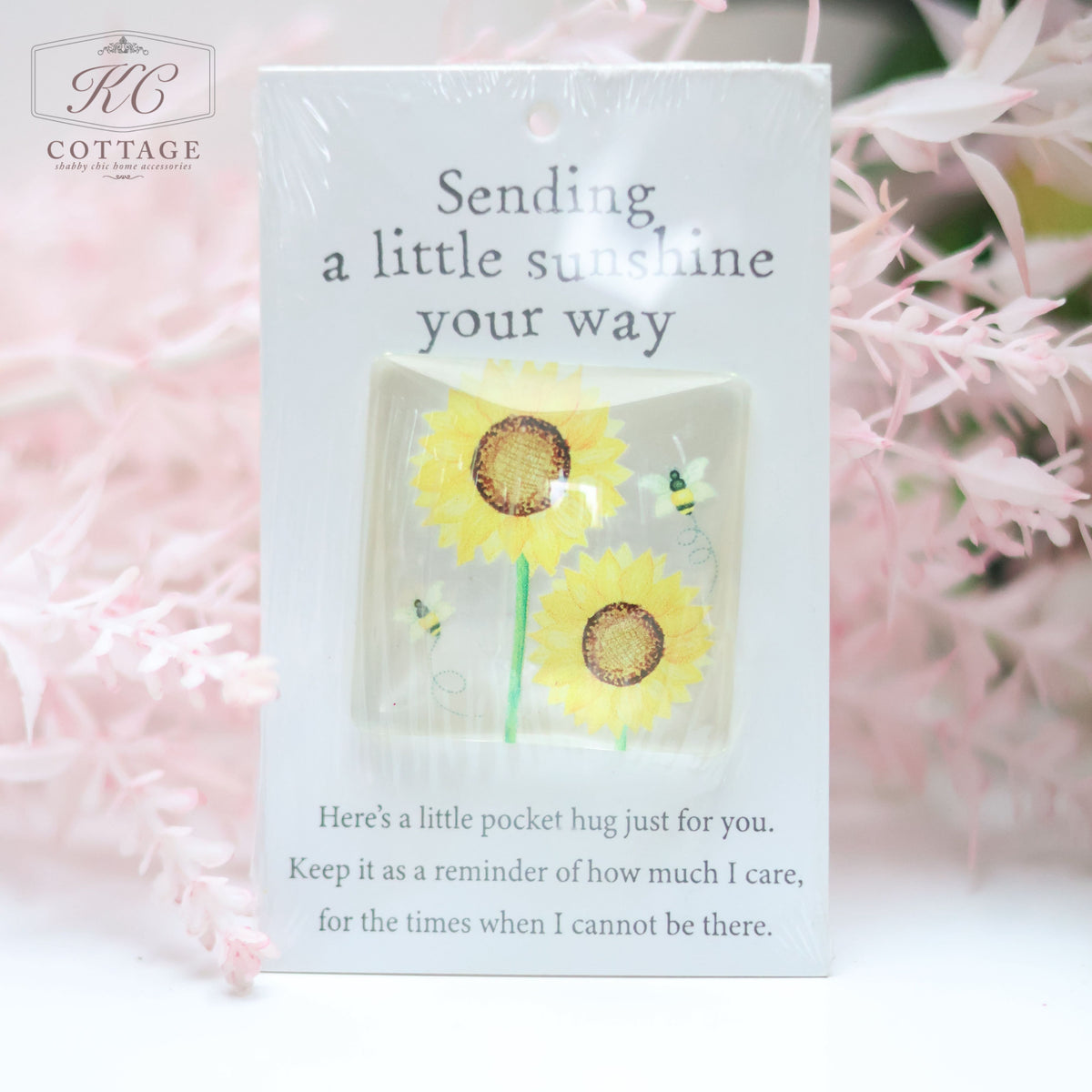 A plastic card with an illustration of two sunflowers and two bees, accompanied by the text "Sending a little sunshine your way." Below, in smaller text: "Here's a little pocket hug just for you. Keep it as a reminder of how much I care, for the times when I cannot be there." The Pocket Hugs with Sentiments card is surrounded by home decor featuring soft, pink foliage.