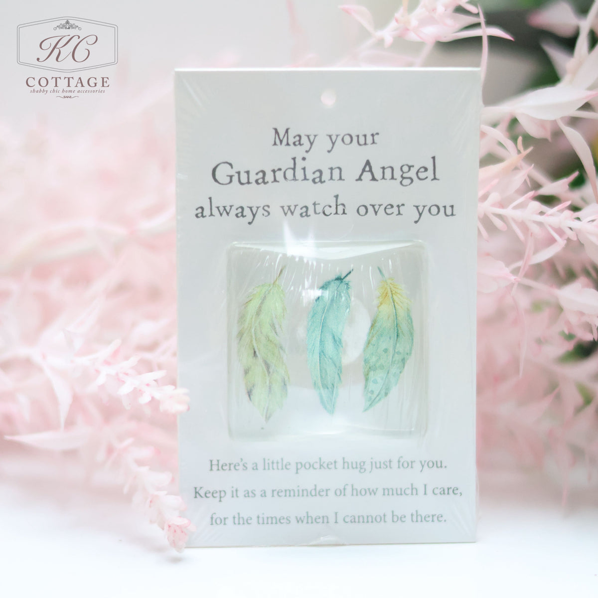 A charming Pocket Hugs with Sentiments with three pastel feather charms. Text reads: "May your Guardian Angel always watch over you. Here’s a little pocket hug just for you. Keep it as a reminder of how much I care, for the times when I cannot be there." Background features pink foliage, perfect for home decor.