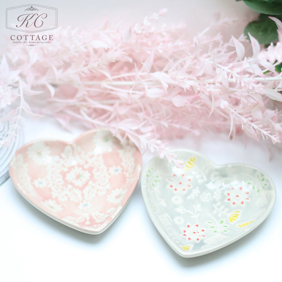 Two **Pastel Hand Painted Floral Heart Trinket Dishes** adorned with intricate floral patterns in pastel pink and light grey hues are placed side by side. They are set against a soft, pinkish-white floral background, enhancing the delicate, cottage-style aesthetic of any home decor.