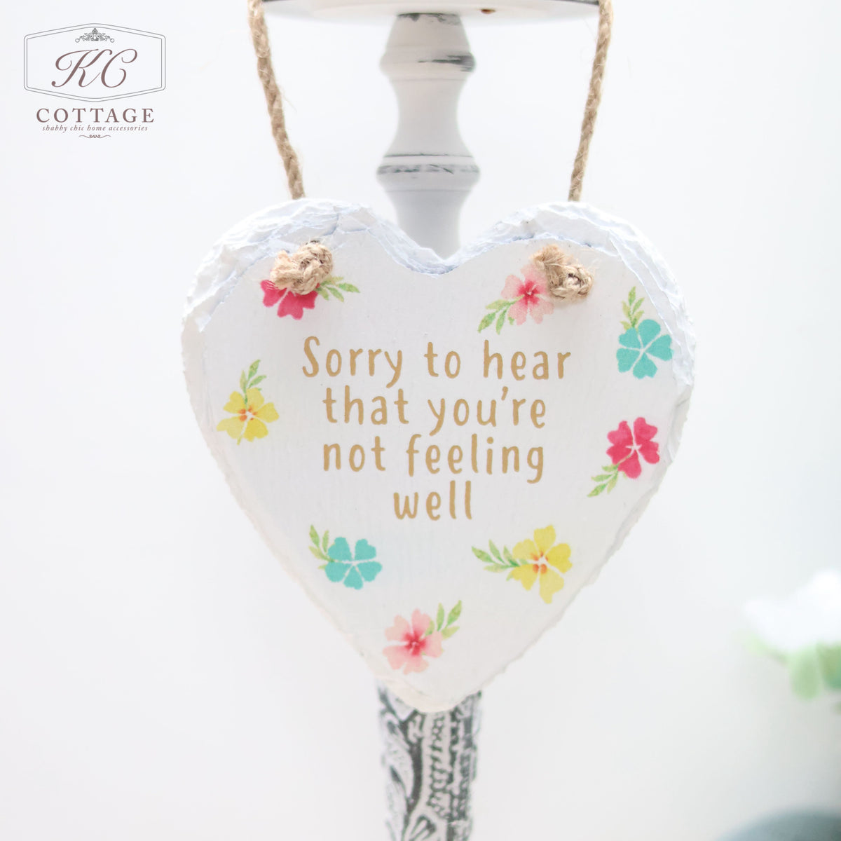 A Bright Floral Slate Hanging Hearts home decor piece with a white background and floral accents. The text on the heart reads, "Sorry to hear that you're not feeling well." It is hanging by a twine string against a light-colored background.