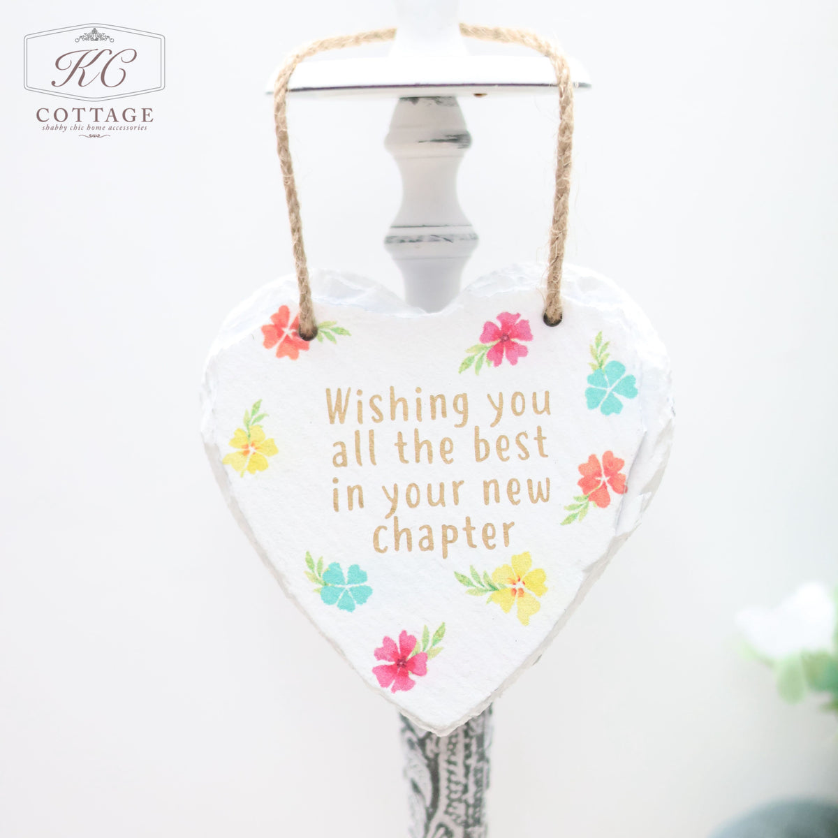 A Bright Floral Slate Hanging Hearts with a rope for hanging features colorful flowers and the message: "Wishing you all the best in your new chapter." The piece of home decor hangs from a white hook, with the background softly out of focus.