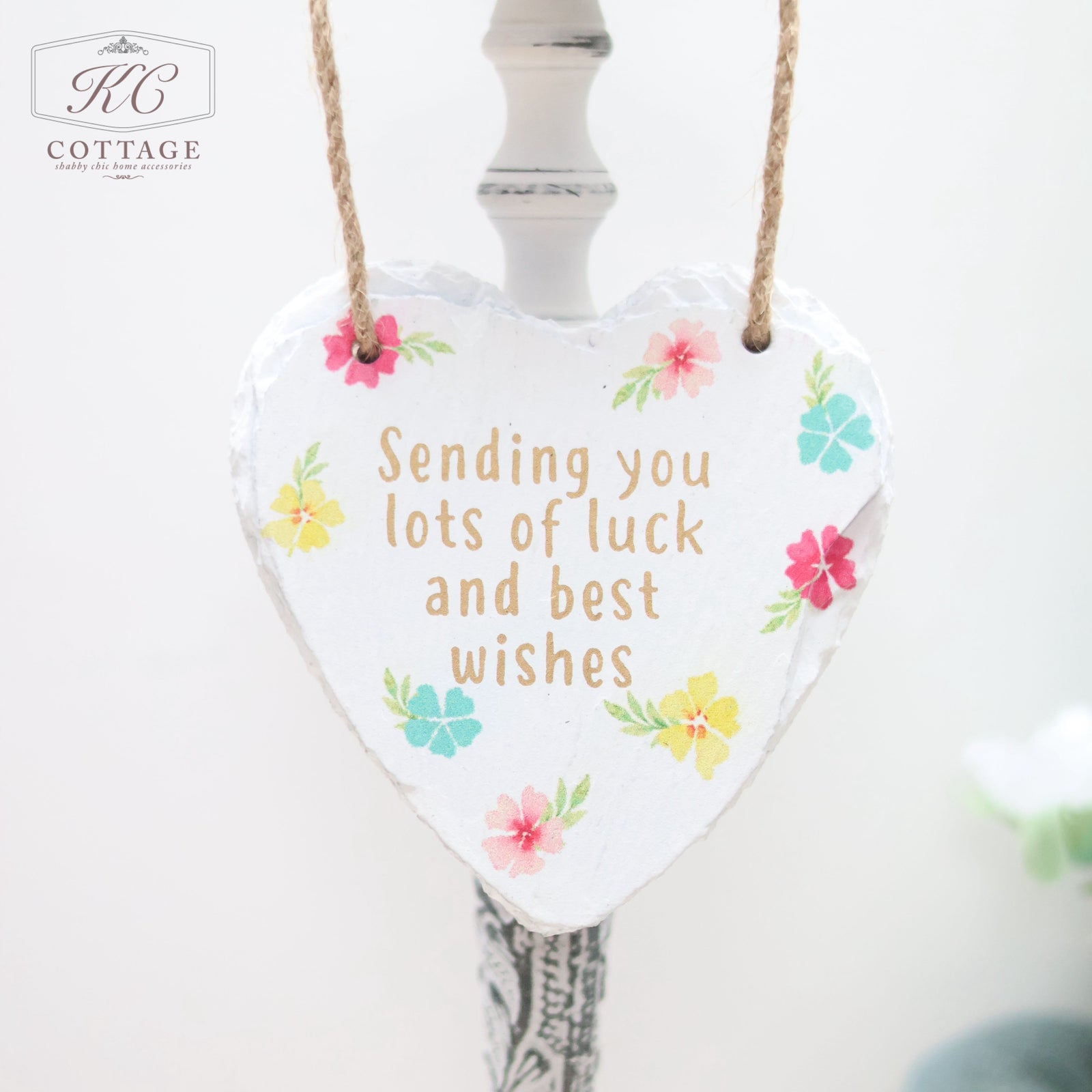 A heart-shaped ornament made of a white material hangs from a brown string. The home decor piece is adorned with colorful flowers and bears the message: "Sending you lots of luck and best wishes." The background is white with a partial view of a light fixture. This charming item is called Bright Floral Slate Hanging Hearts.
