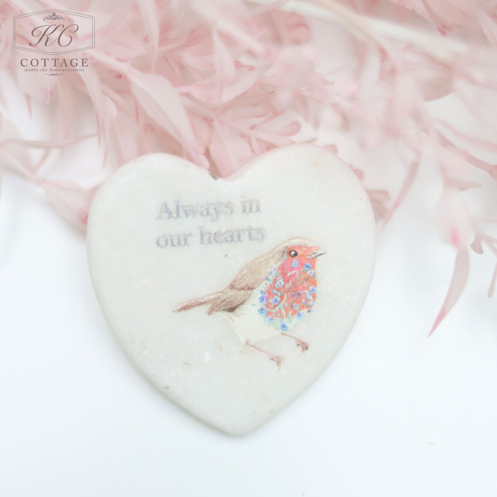 A Marble Pocket Pebble Robin Sentiment Heart ornament featuring an illustration of a small bird and the text "Always in our hearts" is set against a soft, pale pink background with delicate, wispy foliage. Ideal for adding a touch of charm to your home decor.
