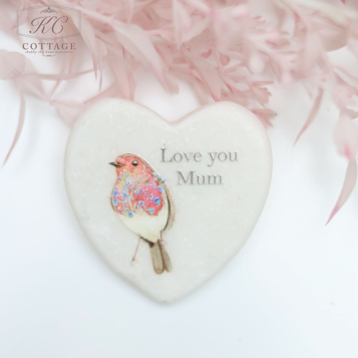 A Marble Pocket Pebble Robin Sentiment Heart with a pastel-colored bird illustration and the text "Love you Mum" is surrounded by soft pink foliage. The background is blurred, highlighting the delicate details of this charming piece of home decor.
