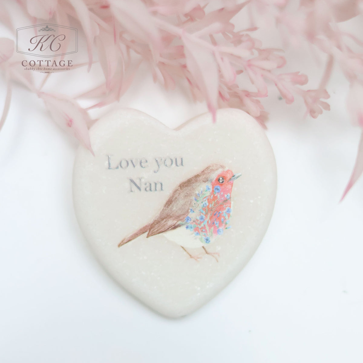 A Marble Pocket Pebble Robin Sentiment Heart with an illustration of a bird and the text "Love you Nan" on it. It is surrounded by soft pink foliage, making it a charming piece of home decor.
