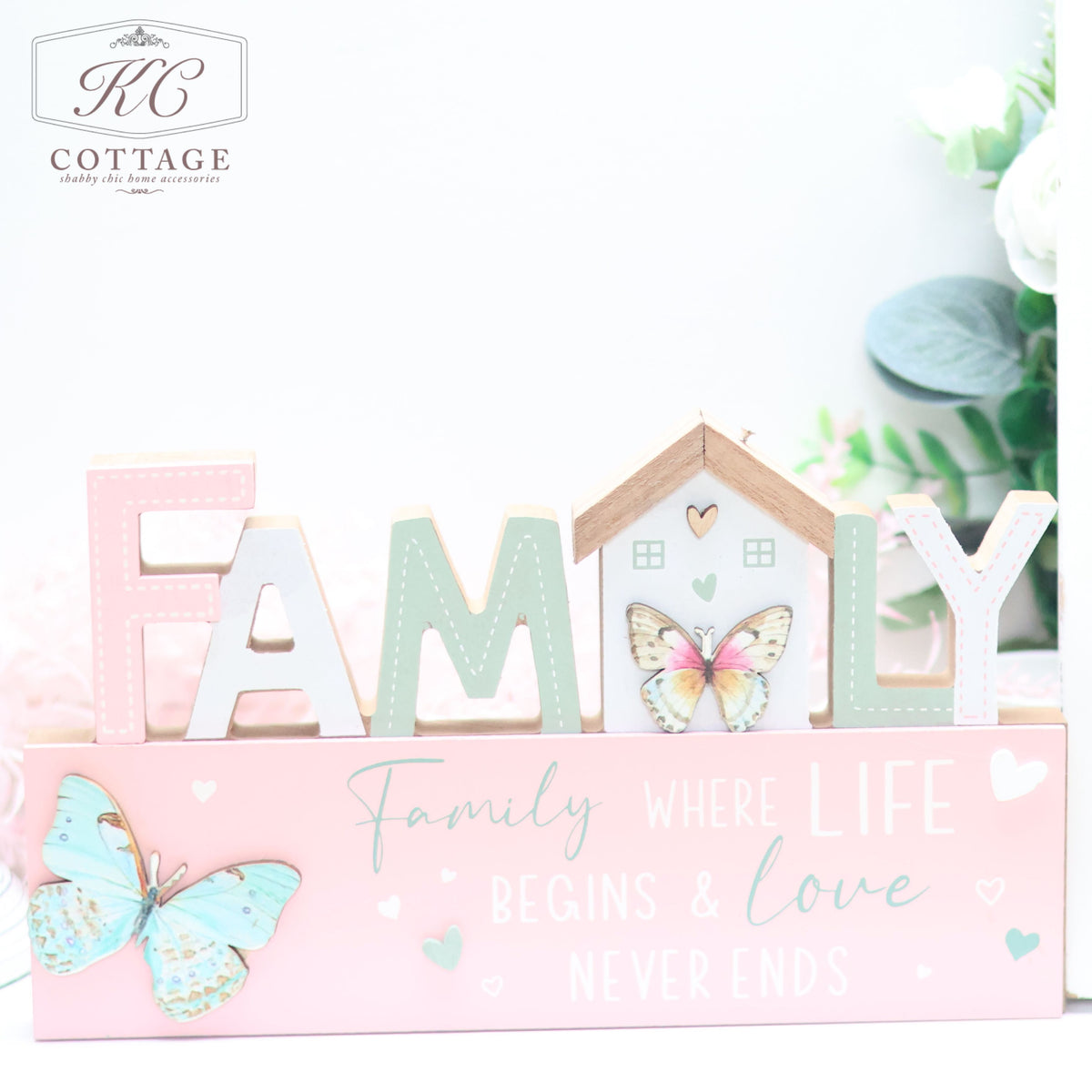A decorative Bright Butterfly Wooden Signs - Family, Friend, Home with pastel colors features the word "FAMILY," where the letter "A" is in pink, "M" is in green, and the "I" is shaped like a house. Below it, a pink base reads: "Family where LIFE begins & love NEVER ENDS." Two butterflies and small hearts adorn this charming piece.