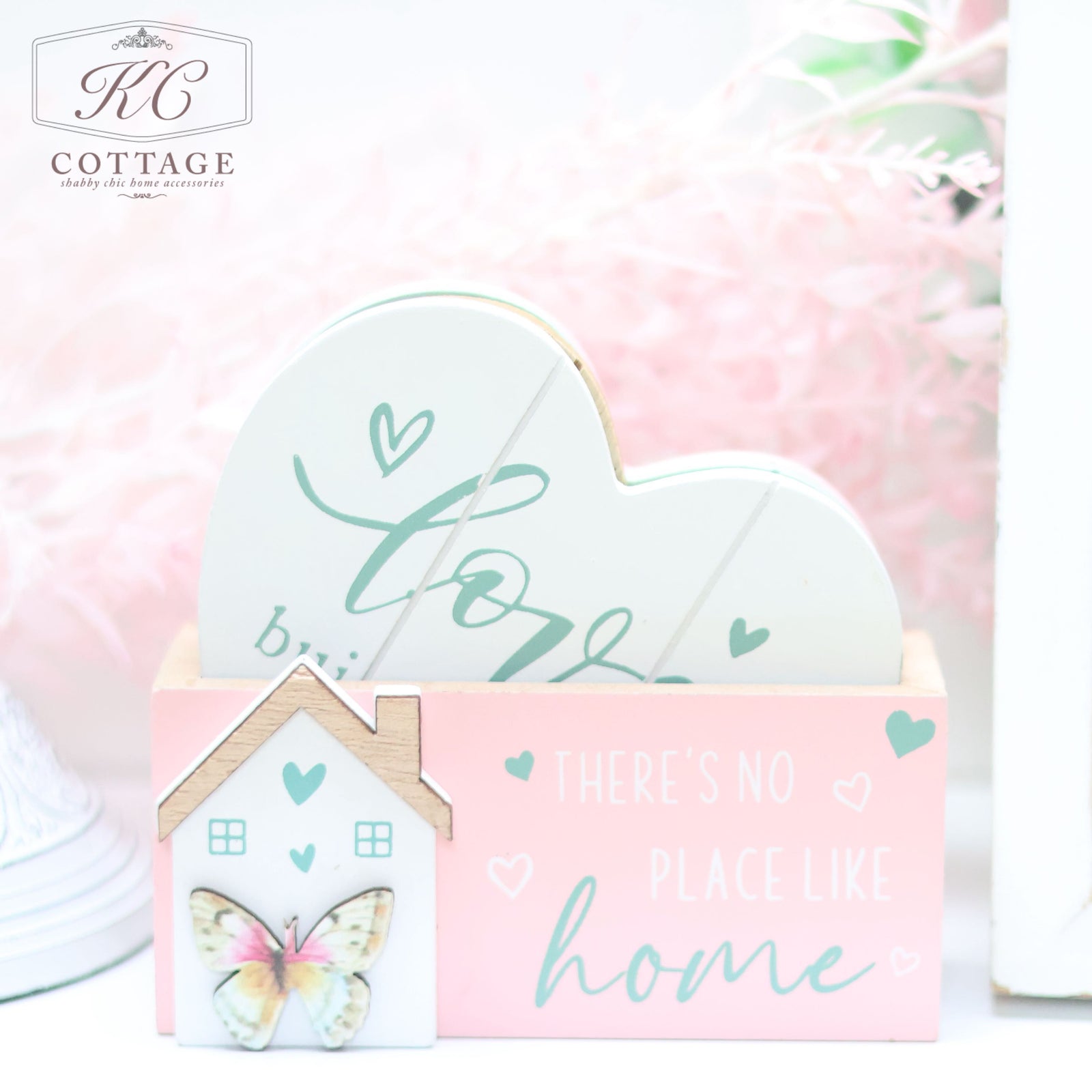 A pastel-colored decorative item featuring a heart-shaped cutout that reads "love" held in a holder. The holder displays a small house with a butterfly illustration and the phrase "There's No Place Like Home." This charming piece adds a touch of elegant home decor to any room can be complemented by the Bright Butterfly Wooden Coasters Set.
