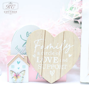 A decorative setup featuring a wooden heart with the text "Family, a circle of love and support" in white. Behind it is a Bright Butterfly Wooden Coasters Set, and other home decor elements. The setup includes a soft, pastel color palette.