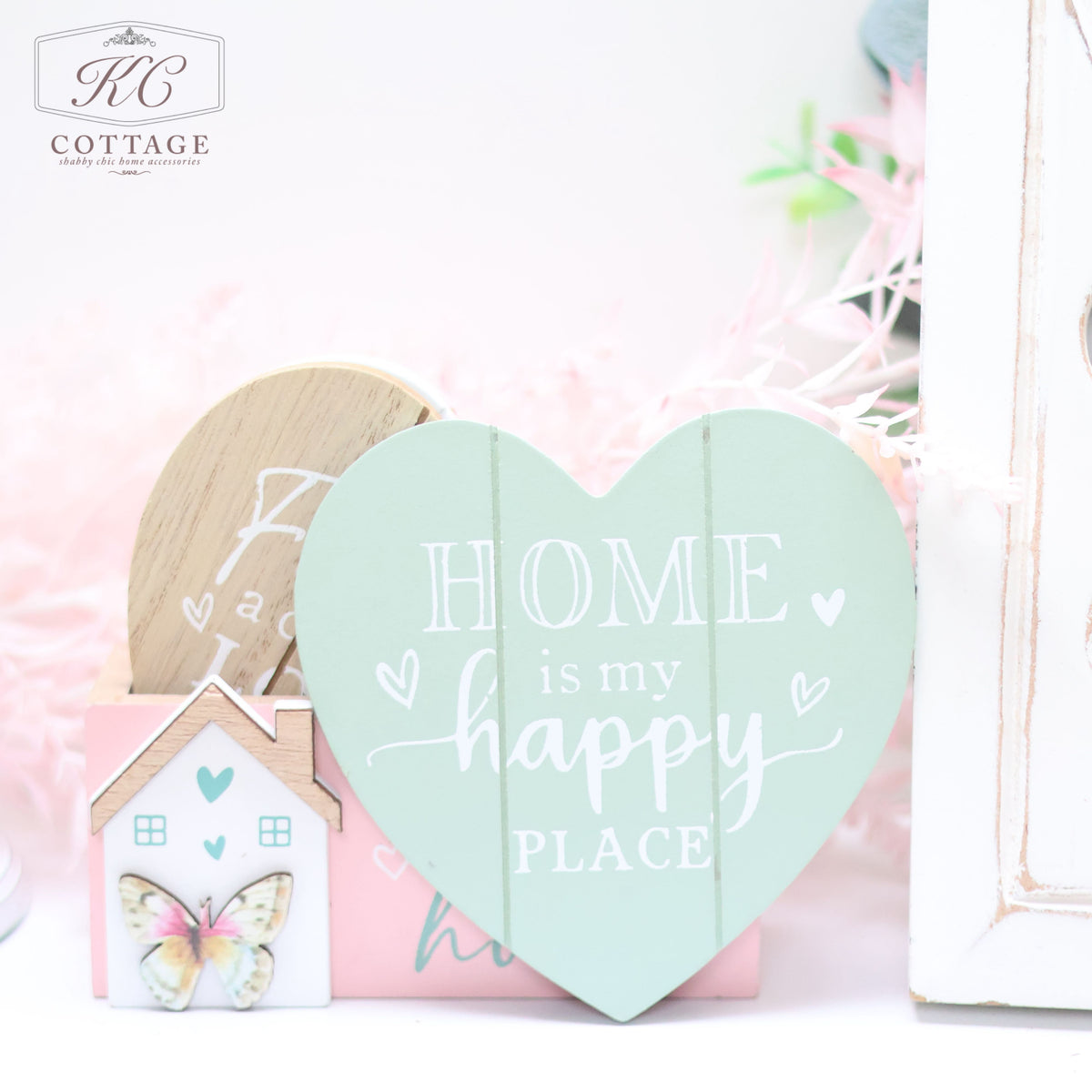 A decorative setup featuring a mint green heart-shaped sign with the words "HOME is my happy PLACE" in white text, next to a Bright Butterfly Wooden Coasters Set. The arrangement includes soft pink, white, and wood accents, perfect for adding charm to your home decor.