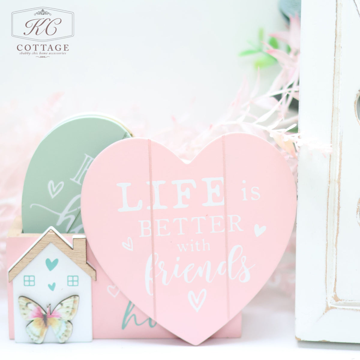 A decorative arrangement featuring a heart-shaped pastel pink sign with the text "Life is better with friends," a pastel green heart, a Bright Butterfly Wooden Coasters Set, and a white background with subtle, soft greenery creates charming home decor.