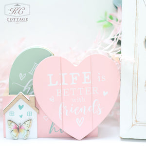 A decorative arrangement featuring a heart-shaped pastel pink sign with the text "Life is better with friends," a pastel green heart, a Bright Butterfly Wooden Coasters Set, and a white background with subtle, soft greenery creates charming home decor.