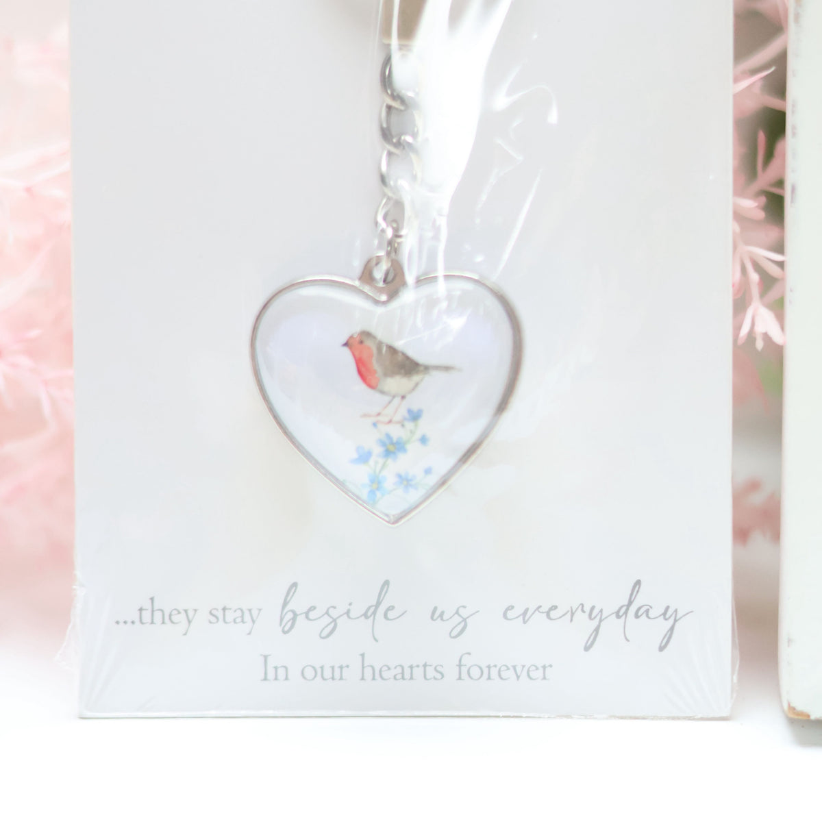 A Glass Forget Me Not Heart Keyring featuring an illustration of a bird, presented against a white background. Perfect for home decor, the packaging has the text: "...they stay beside us every day. In our hearts forever" written in cursive at the bottom.