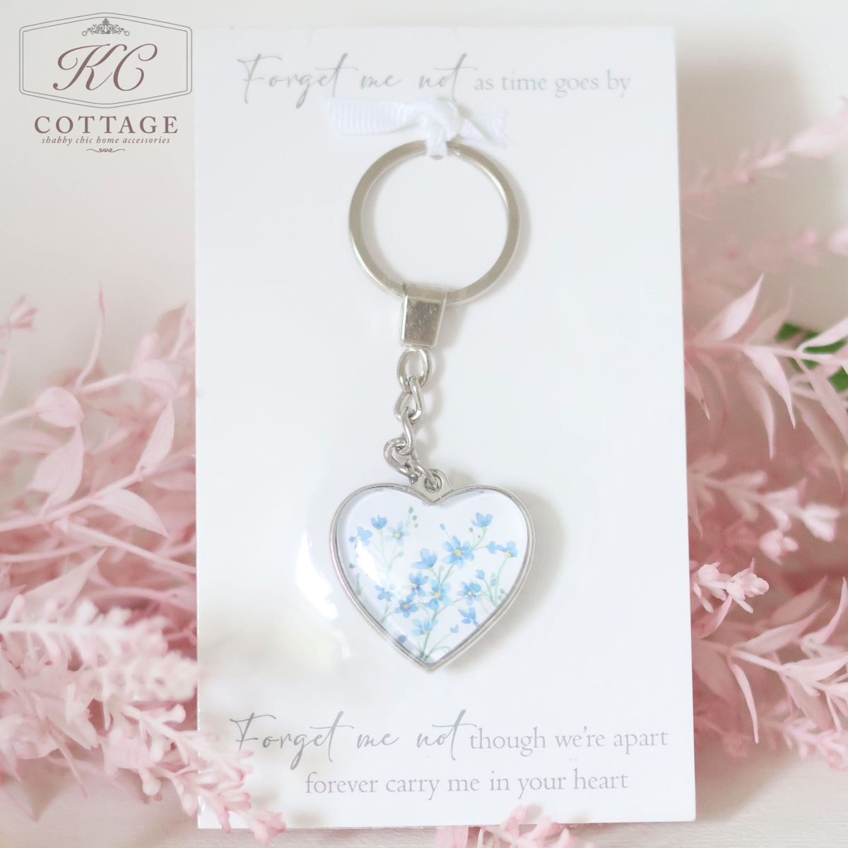 A Glass Forget Me Not Heart Keyring is displayed against a white card with text. The card reads, "Forget me not as time goes by" and "Forget me not though we’re apart, forever carry me in your heart." Pink flowers are in the background, complementing home decor accents.
