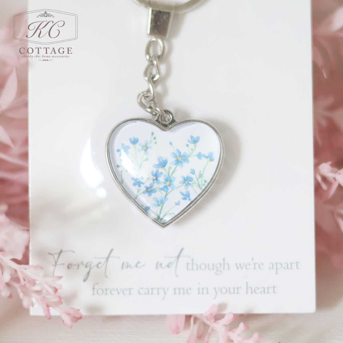 A Glass Forget Me Not Heart Keyring featuring delicate blue forget-me-not flowers is attached to a card with the message: "Forget me not, though we're apart, forever carry me in your heart." Perfect as a piece of home decor, the card and keyring are set against a soft pink, floral background.