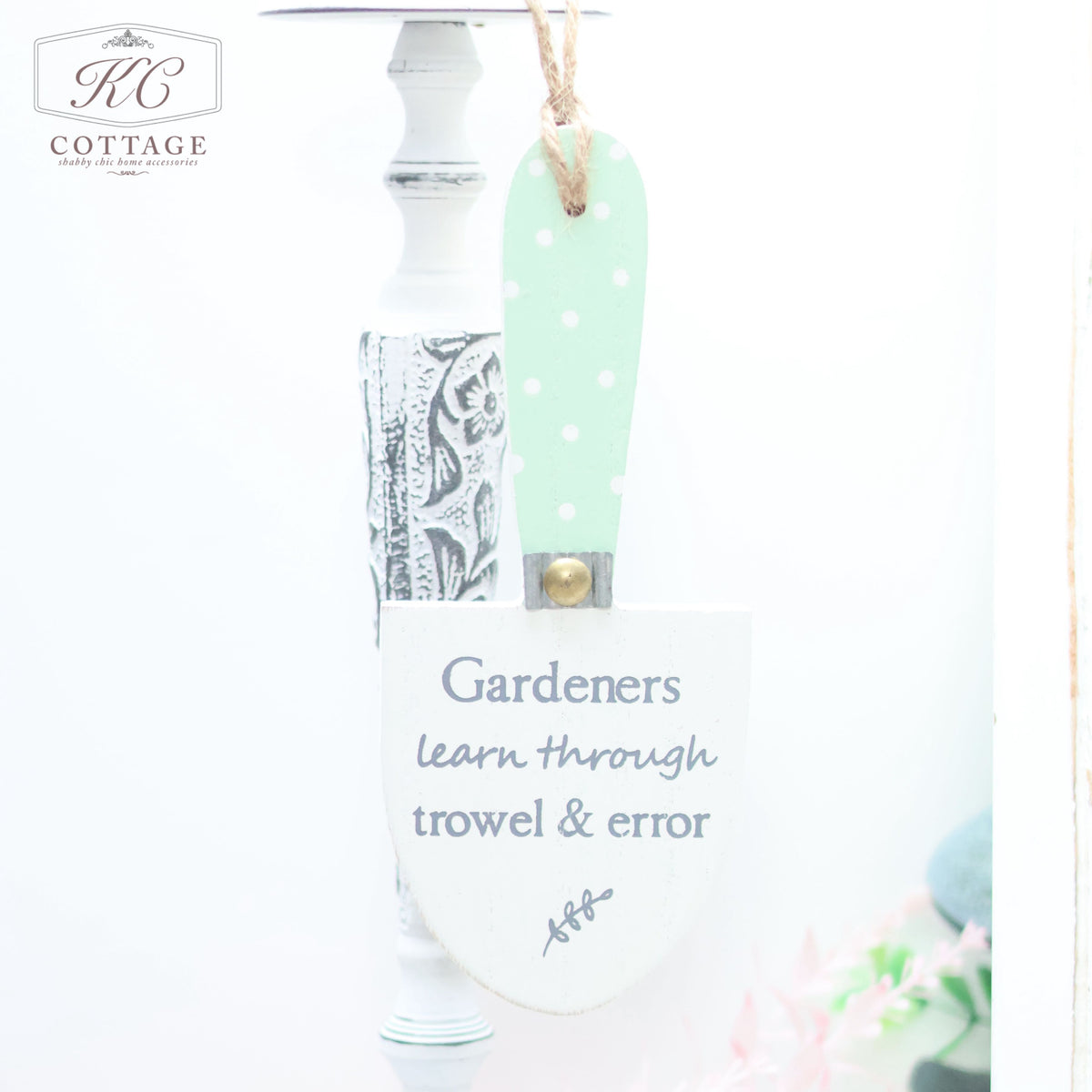 A decorative white and green Wooden Garden Trowel Hanger with a polka dot pattern hangs from a white spindle, perfect for adding charm to your home decor. The trowel bears the text, "Gardeners learn through trowel & error." A faint glimpse of pink and green elements graces the background. The KC Cottage logo is at the top left.