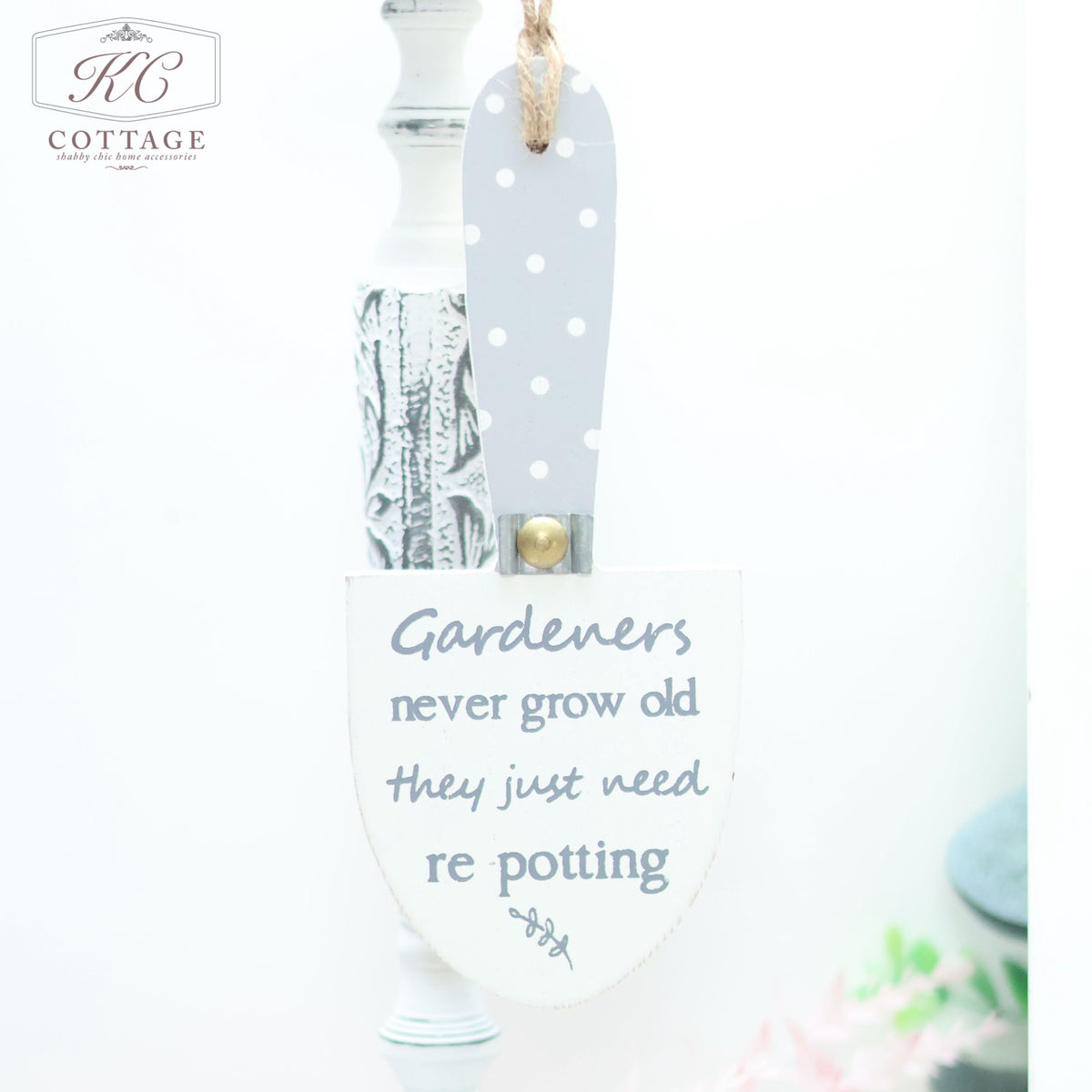 A Wooden Garden Trowel Hanger shaped like a small garden trowel hangs on a white, ornate background. The home decor piece is white with gray polka dots and has the text, "Gardeners never grow old, they just need re-potting," written in cursive. The KC Cottage logo is in the corner.