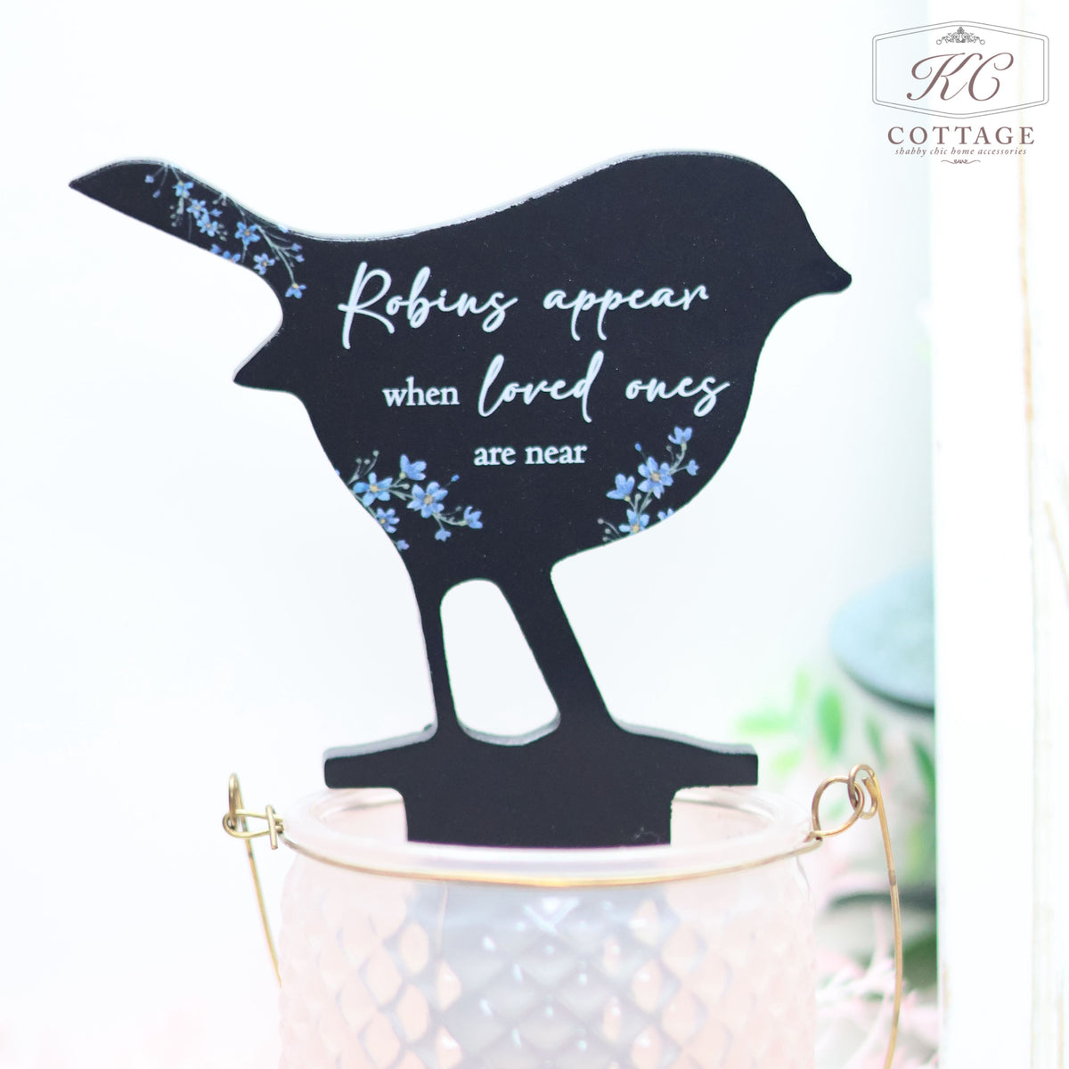 A decorative black bird silhouette with blue floral accents stands on a light pink textured jar. The bird features the text, "Robins appear when loved ones are near." This charming piece makes for perfect home decor. The Wooden Robin Forget Me Not Stake adds a touch of elegance and remembrance to any space.