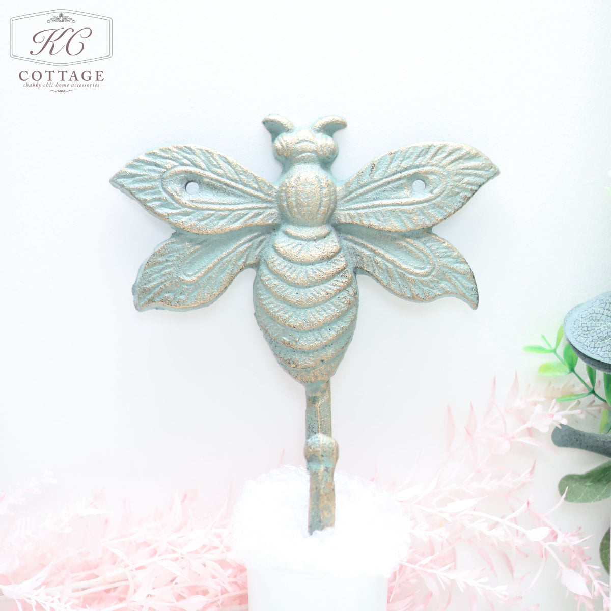 Rustic, Wrought Iron Bee Wall Hook from the KC Cottage brand is mounted on a pale background. The metal hook features detailed wings and body segments, with greenery and pink foliage decor below adding an elegant touch to your home decor.