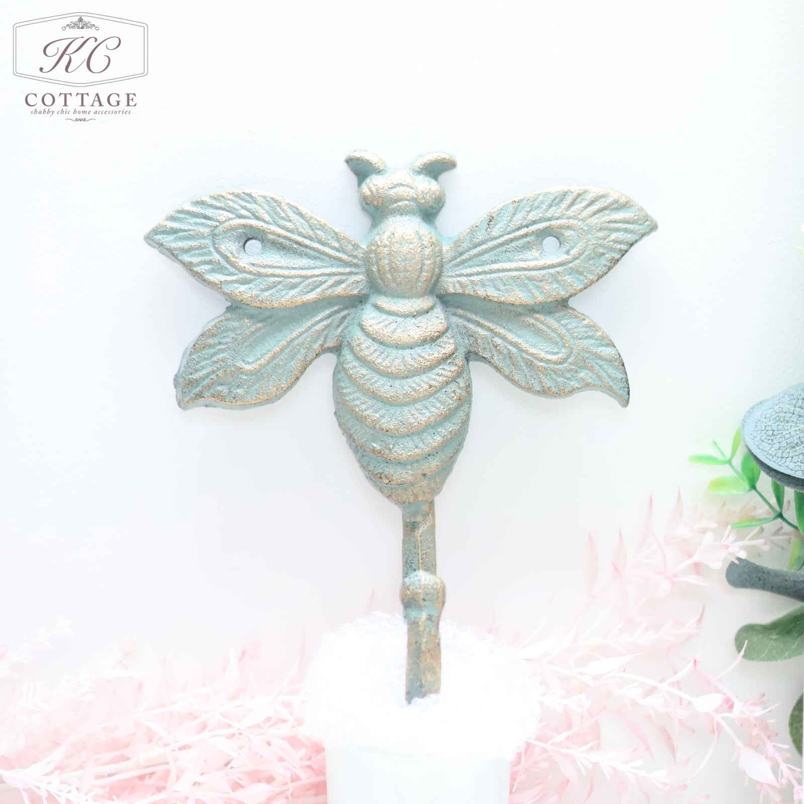 Rustic, Wrought Iron Bee Wall Hook from the KC Cottage brand is mounted on a pale background. The metal hook features detailed wings and body segments, with greenery and pink foliage decor below adding an elegant touch to your home decor.
