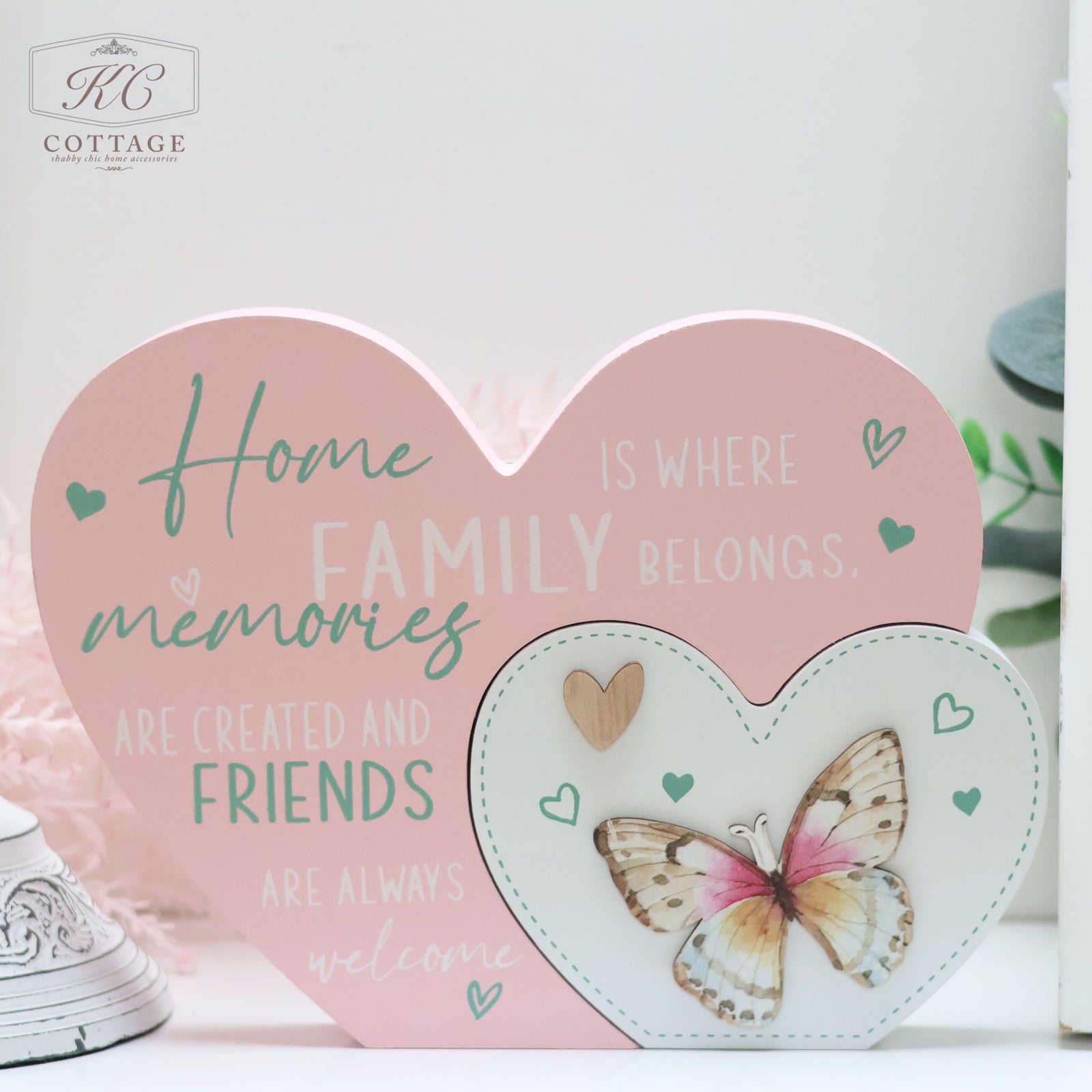 A decorative pink heart-shaped sign with a smaller heart attached, perfect for home decor. The large heart reads, "Home is where family belongs, memories are created and friends are always welcome." The smaller heart features a wooden butterfly with colorful wings and heart motifs around it. Presenting: Bright Butterfly Heart Plaques.
