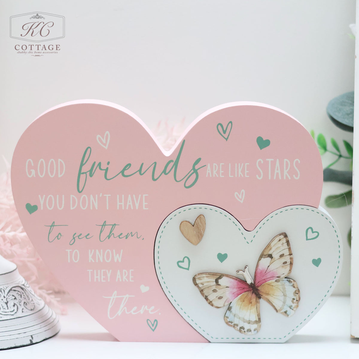 A decorative home decor display by KC Cottage features a larger pink heart with the text "Good friends are like stars. You don't have to see them to know they are there." Next to it is a smaller heart-shaped frame with a butterfly image inside, collectively known as Bright Butterfly Heart Plaques.