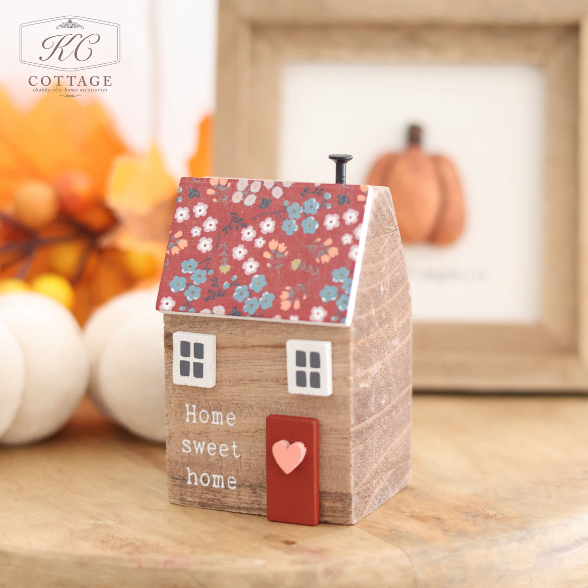 Introducing the Wooden House With Floral Roof: a charming small wooden house figurine featuring a floral-patterned roof and a red door adorned with a heart. The front of the house is beautifully inscribed with the words "Home sweet home." Complementing this delightful ornament are fall decorations, including white pumpkins and a framed picture of a pumpkin, making it an ideal addition to your autumn decor.