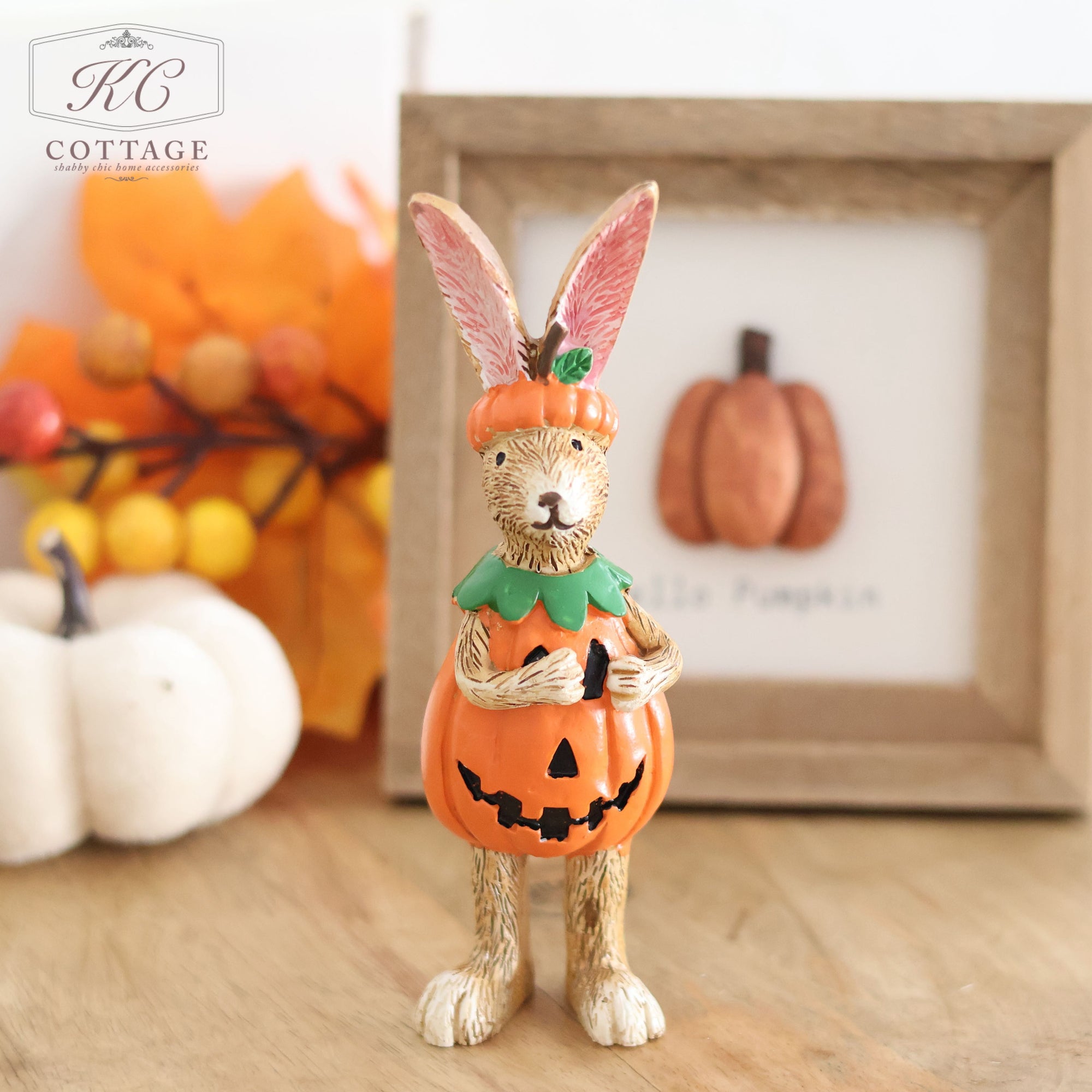 An Autumn Standing Pumpkin Rabbit figurine, dressed adorably in a pumpkin costume, is placed on a wooden surface. In the background, there's a framed picture of a pumpkin, surrounded by small white pumpkins and textured autumn leaves. The text "KC Cottage" is displayed in the top left corner.