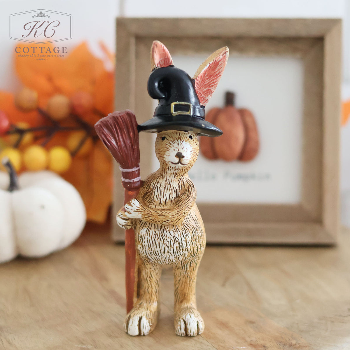The Halloween Rabbit Standing Decoration with Broom & Hat features a charming rabbit in a black witch hat holding a broom, positioned on a wooden surface. In the background, there's a framed picture of pumpkins, gourds, and fall leaves, creating the perfect Halloween atmosphere. This spooky home decor is branded "KC Cottage.