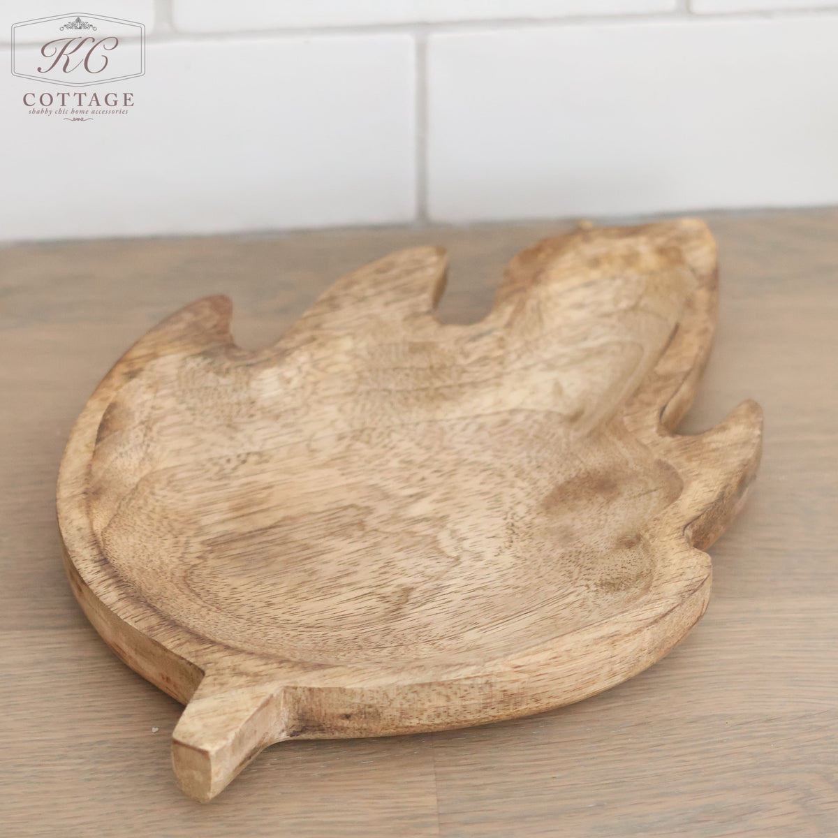 A Mango Wood Leaf Tray, crafted with a leaf design, is displayed on a wooden surface against a backdrop of white tiles. The tray showcases a natural wood finish and detailed grain patterns, radiating organic elegance. In the upper left corner, the "KC Cottage" logo is visible. Ideal for home decor enthusiasts.