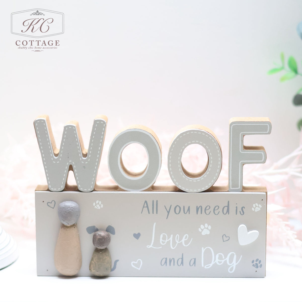 A decorative wooden "Dog Pebble Sign" features the large word "WOOF," accompanied by two charming figurines—a person and a dog. Beneath it, the sign reads, "All you need is Love and a Dog," accentuated with small paw prints, hearts, and the logo "KC Cottage." Perfect for any dog lover's home, the background is softly blurred.