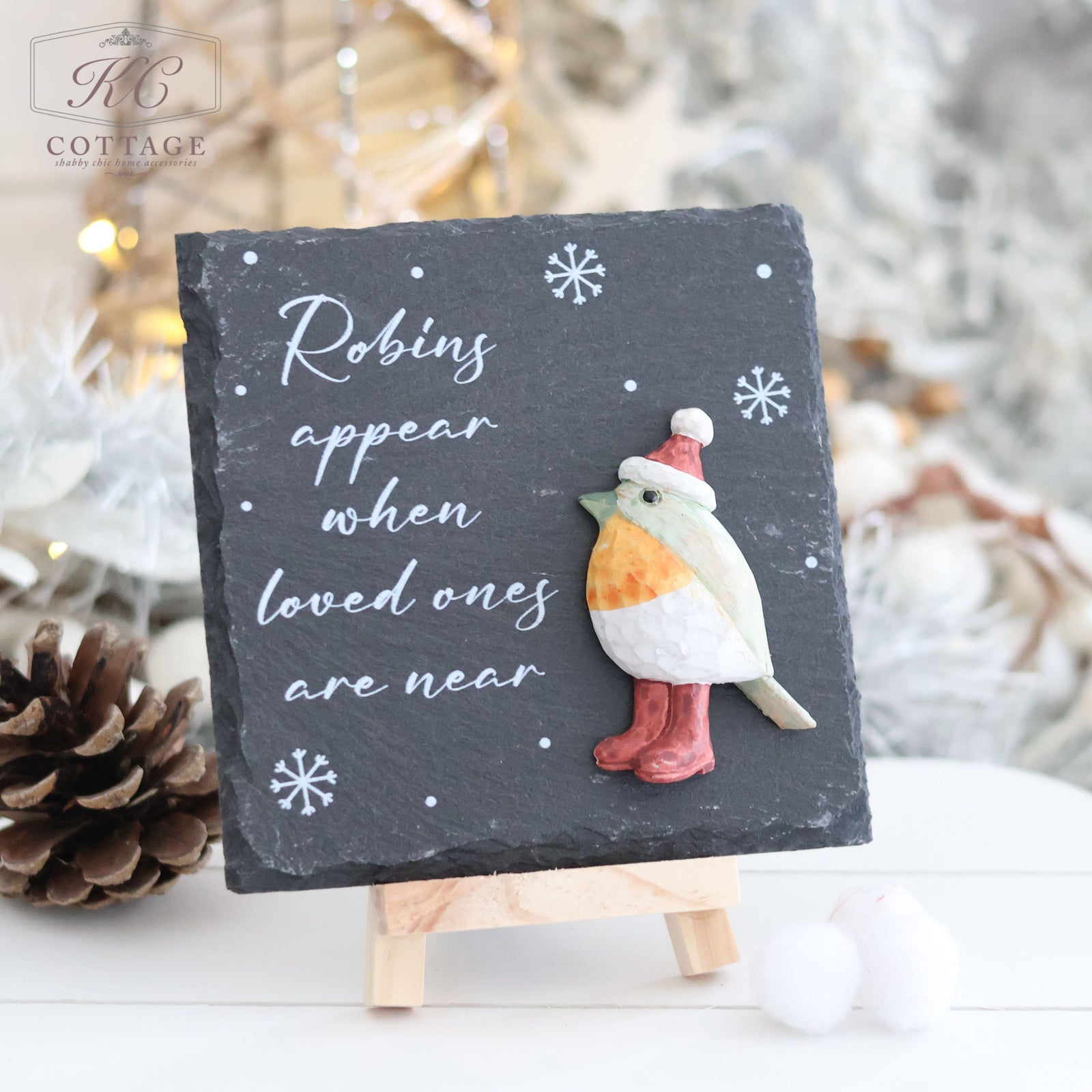 The Christmas Slate Robins Appear Easel Plaque is a charming small black slate sign on a wooden stand that features the text "Robins appear when loved ones are near" in white cursive. Adorned with a clay robin wearing a pink hat and red boots, the plaque is set against a background brimming with pinecones and holiday cheer.