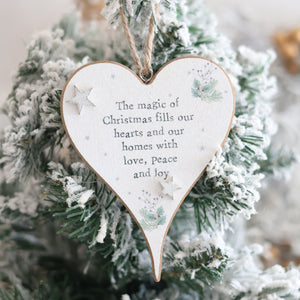 Hanging on a snow-covered tree branch, the Christmas Sentiment Heart Hanger With 3D Star features the text: "The magic of Christmas fills our hearts and our homes with love, peace and joy." White stars and festive decorations embellish the ornament, adding to the festive joy.