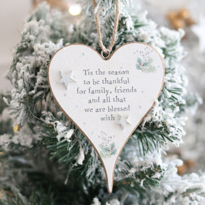The Christmas Sentiment Heart Hanger With 3D Star adorns a snow-covered Christmas tree, exuding festive joy. Featuring 3D star decorations and the inscription, "Tis the season to be thankful for family, friends, and all that we are blessed with," this ornament is completed with small festive illustrations.