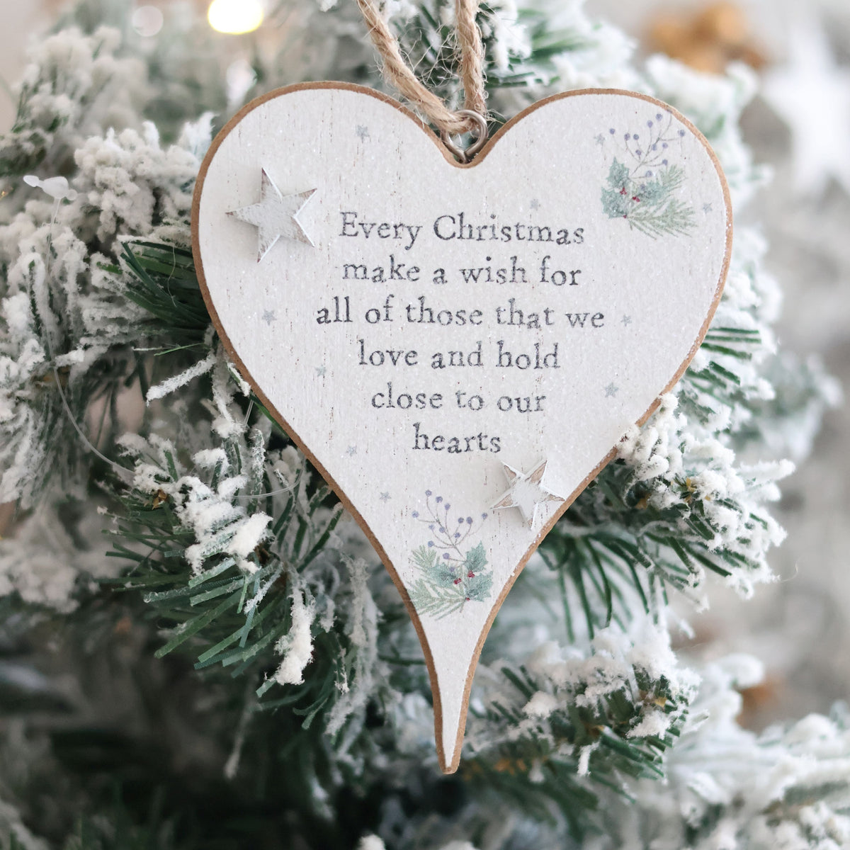 The Christmas Sentiment Heart Hanger With 3D Star, adorned with the inscription "Every Christmas make a wish for all of those that we love and hold close to our hearts," hangs on a snow-dusted Christmas tree. Decorated with small stars and pine branches, it radiates festive joy.