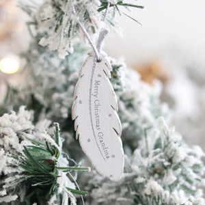 A Christmas Family Sentimental Feather Hanger hangs on a snowy Christmas tree branch. The ornament, a cherished memory, has the words "Merry Christmas, Grandma" inscribed on it in delicate text, surrounded by small star designs. The background is softly blurred with festive lights.
