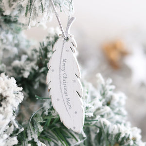 A delicate white feather-shaped ornament, the Christmas Family Sentimental Feather Hanger, hangs from a frosted Christmas tree. Inscribed with "Merry Christmas Mum" in elegant script and adorned with small silver stars, this cherished memory adds a touch of magic as snow-like frost covers the surrounding pine needles.